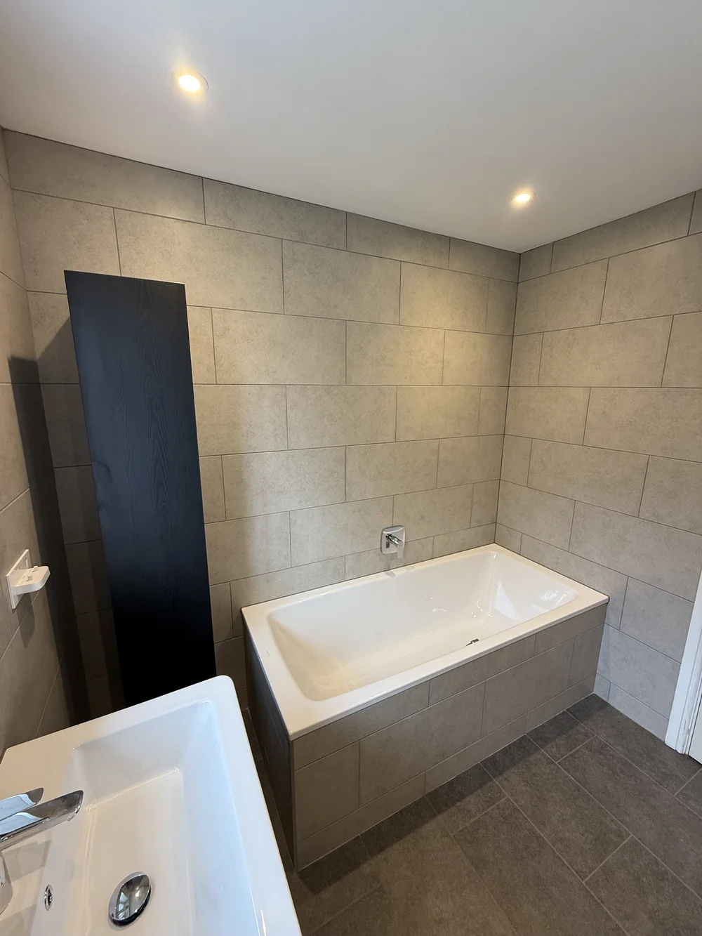 Stone-tiled bathroom with panelled bath and tall black storage column — bathroom renovation Godalming