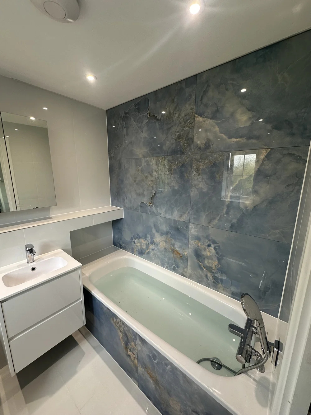 Family bathroom with blue onyx-effect feature wall, panelled bath and floating white vanity — bathroom installation Godalming
