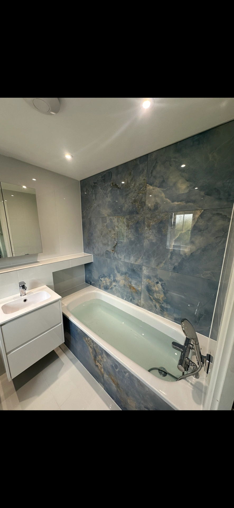 Bathroom with blue onyx-effect feature wall, panelled bath and floating white vanity — bathroom renovation Godalming
